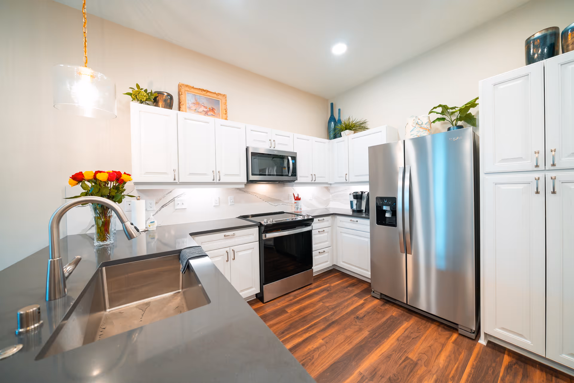 Modern kitchen with white cabinets, stainless steel refrigerator, oven, microwave, and a sink with a curved faucet. There are decorative plants and a vase with red and yellow roses on the countertop. The floor is wooden and the lighting is bright.