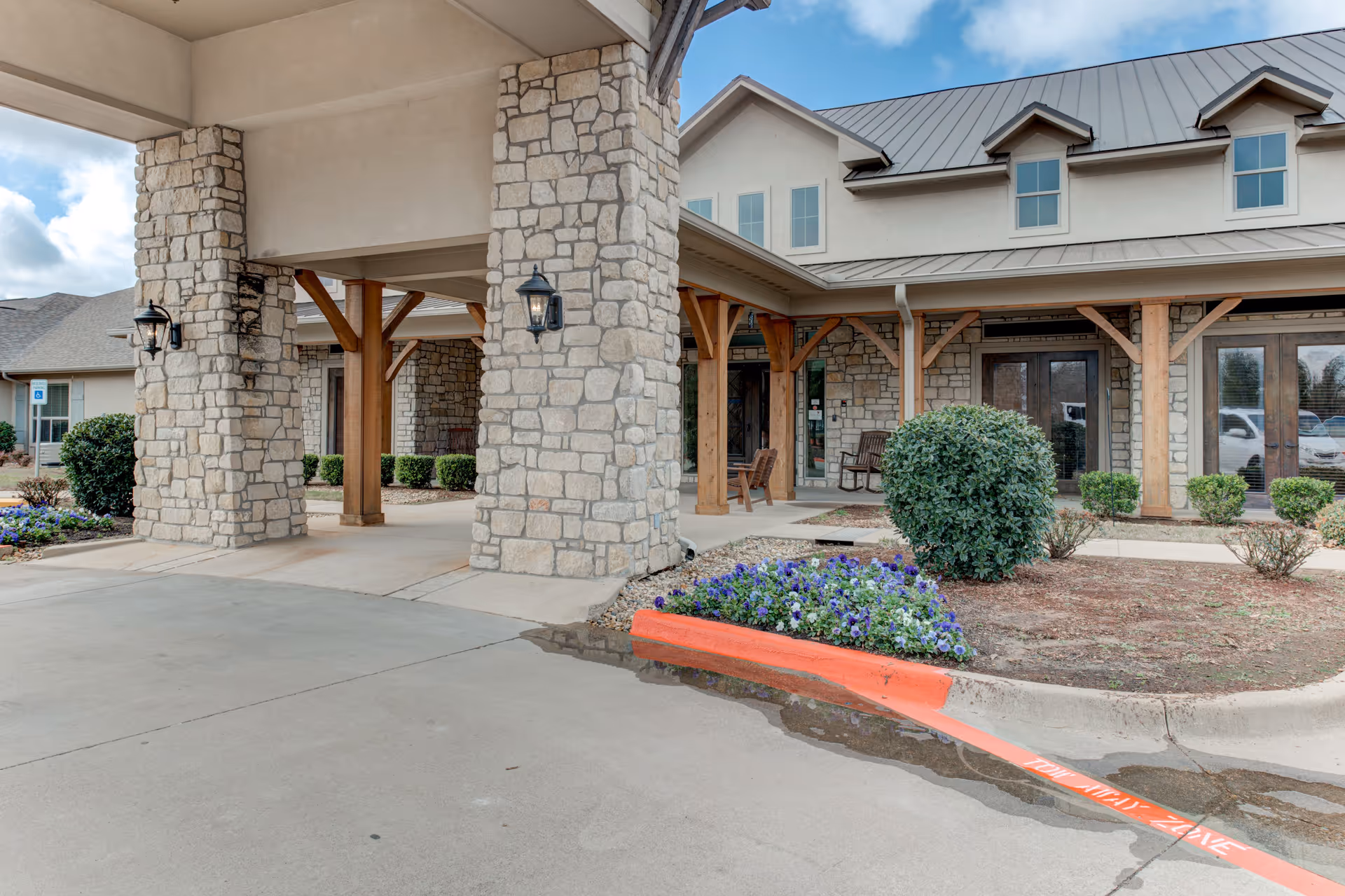 Entrance area of a senior living facility with stone pillars, wooden beams, and a covered driveway. There are bushes, flowers, and a building with multiple windows and a metal roof in the background.