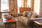 A cozy living room with a brown leather sofa adorned with patterned cushions, a wooden coffee table decorated with a fall-themed centerpiece, a wooden side table, and a wooden dresser with a lamp and decorative items. Large windows with floral curtains allow natural light to fill the room.