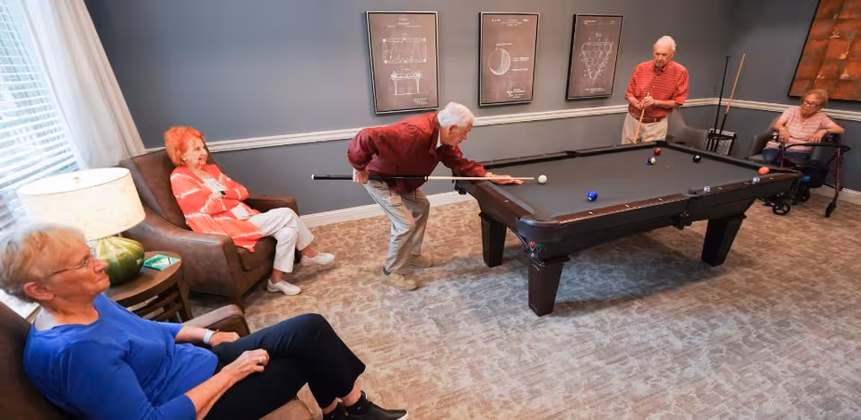 A group of elderly people in a room playing pool. One man is leaning over the pool table aiming a shot, another man is standing holding a cue stick, and two women are seated nearby watching the game. The room has gray walls with framed diagrams of billiard balls and cues, a carpeted floor, and a window with white curtains letting in natural light.