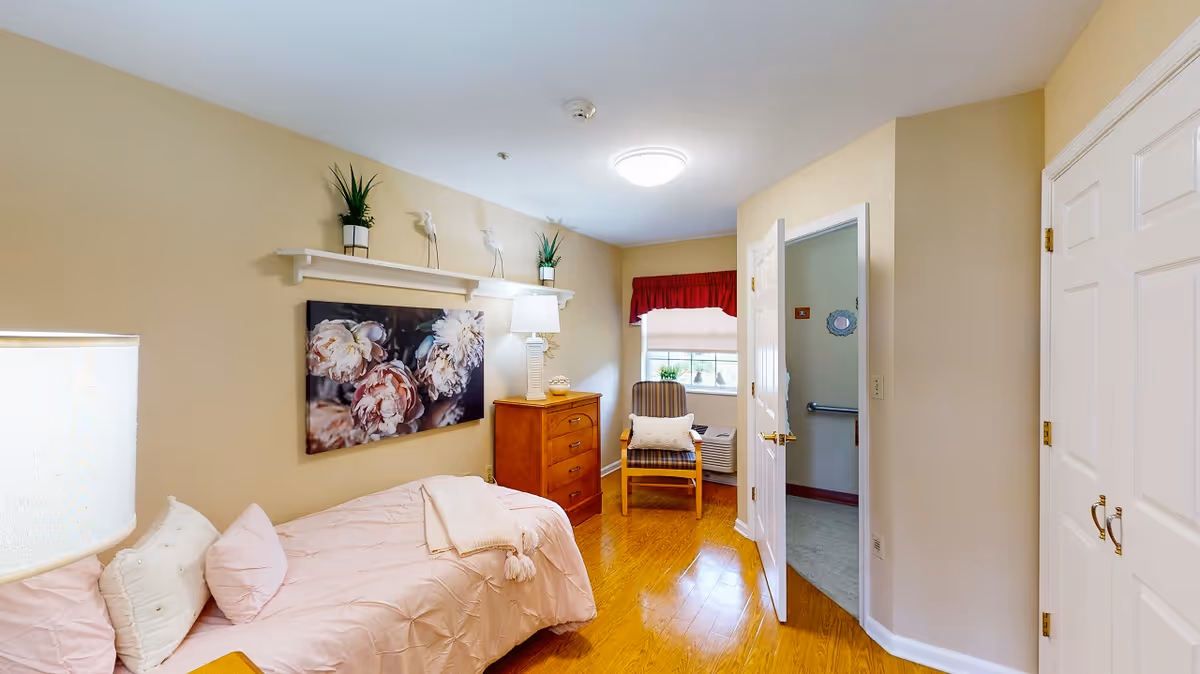 Small, well-lit single bedroom with a twin bed, floral wall art, a wooden dresser and a chair by the window.
