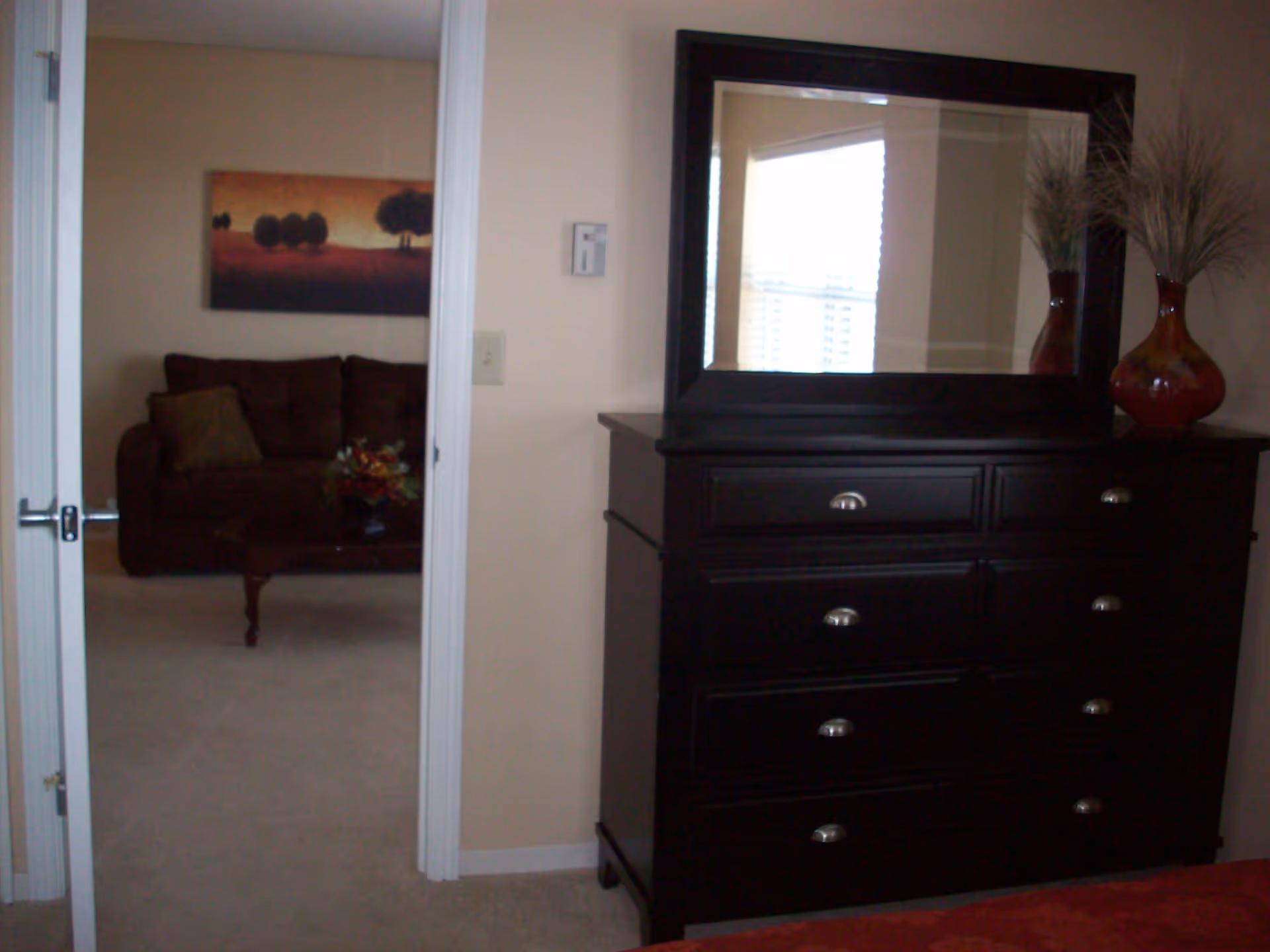 Interior view of a senior living facility room showing a dark wooden dresser with a large mirror on top and a decorative vase with dried plants. Through an open door, a living room area is visible with a dark brown sofa, a coffee table with a flower arrangement, and a landscape painting on the wall.