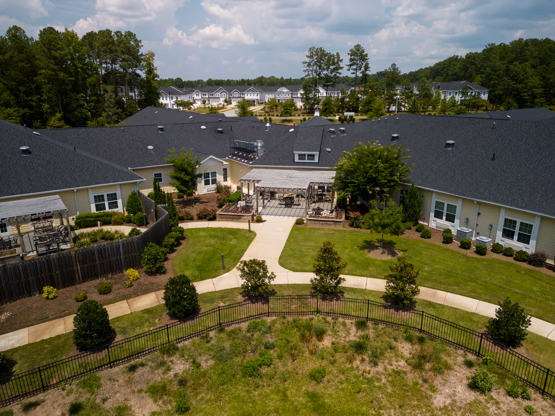 Aerial view of the outdoor courtyard area of Cadence Garner senior living facility, showing a large building with a dark roof surrounding a landscaped garden with pathways, small trees, shrubs, and a pergola-covered seating area. In the background, there are additional residential buildings and a wooded area under a partly cloudy sky.