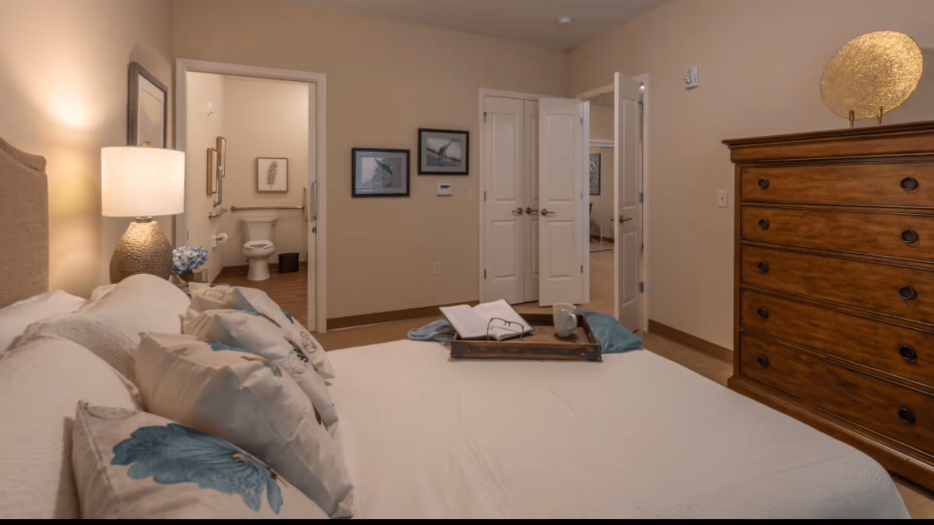 A cozy bedroom with a large bed adorned with multiple pillows and a tray holding a book and a mug. To the left, there is a bedside table with a lamp and a small flower arrangement. The room has beige walls and wooden flooring. In the background, an open door reveals a bathroom with a toilet and framed artwork on the walls. Another set of double doors is partially open, showing a glimpse of an adjacent room.