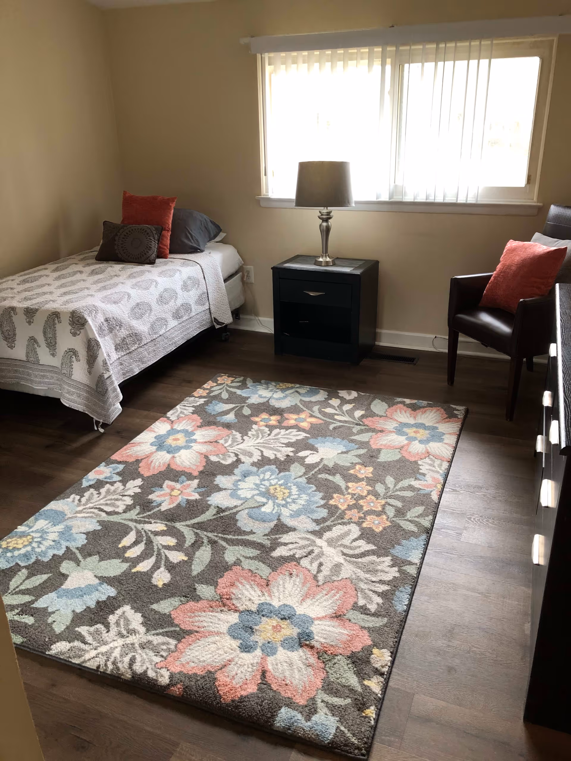 A small bedroom with a single bed covered with a white and gray patterned bedspread and two pillows, one red and one dark gray. Next to the bed is a black nightstand with a silver lamp. There is a window with vertical blinds above the nightstand. A dark brown chair with a red pillow is positioned near the window. The floor is wooden with a large floral area rug in shades of pink, blue, and green.