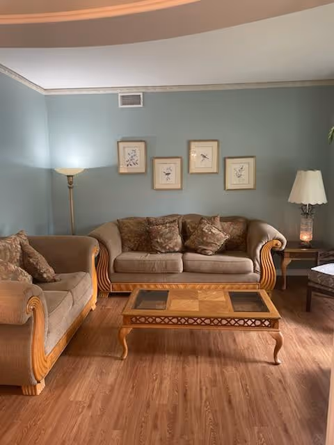 A cozy living room with two beige upholstered sofas adorned with patterned cushions, a wooden coffee table with a glass inset, a floor lamp in the corner, and a side table with a decorative lamp. The walls are painted light blue and decorated with four framed botanical prints.