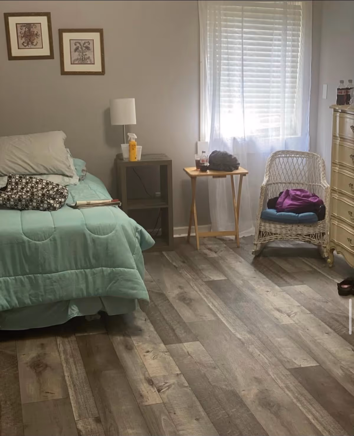 A bedroom with a bed covered in a light green comforter and pillows. Next to the bed is a small nightstand with a lamp and some items on it. There is a small wooden table near a window with sheer white curtains, holding a few items including a drink. A white wicker chair with cushions and clothing on it is positioned next to a cream-colored dresser. The floor is made of wood with a rustic finish. Two framed pictures hang on the wall above the bed.