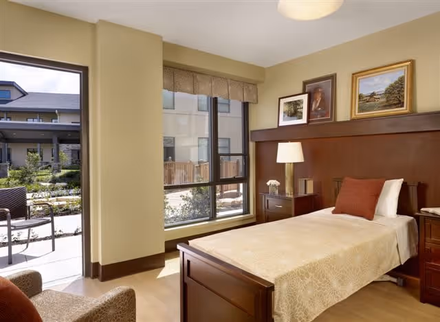 A bright and cozy bedroom with a single wooden bed covered in a beige bedspread and a rust-colored pillow. Next to the bed is a wooden nightstand with a lamp and a small flower vase. The room has large windows letting in natural light and offering a view of an outdoor patio area with chairs and greenery. The walls are decorated with framed pictures above the bed's headboard.