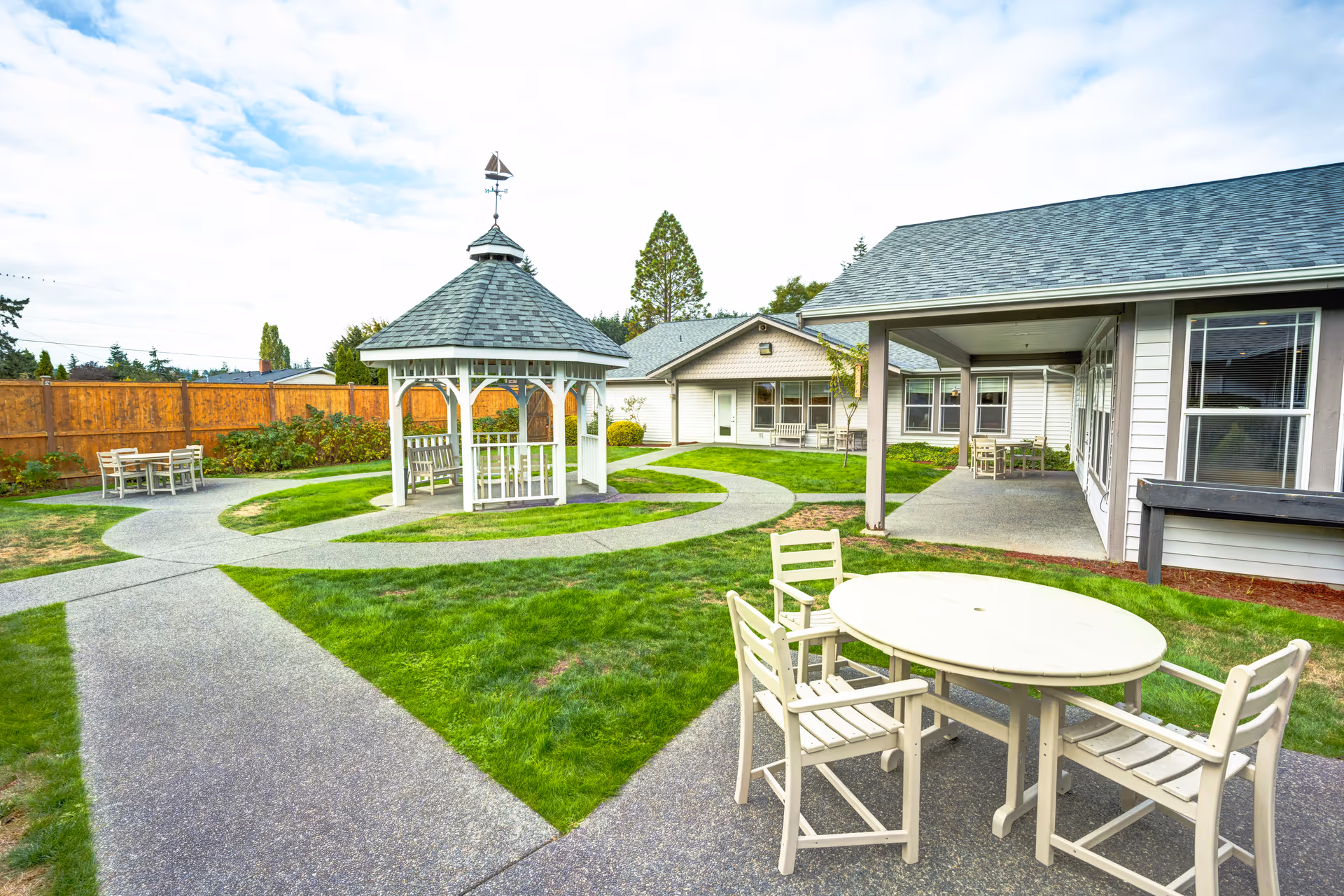 Outdoor courtyard area at HomePlace Special Care at Oak Harbor featuring a white gazebo with a weather vane on top, surrounded by green grass and paved walkways. There are several white wooden tables and chairs placed around the courtyard, with a building visible in the background under a partly cloudy sky.