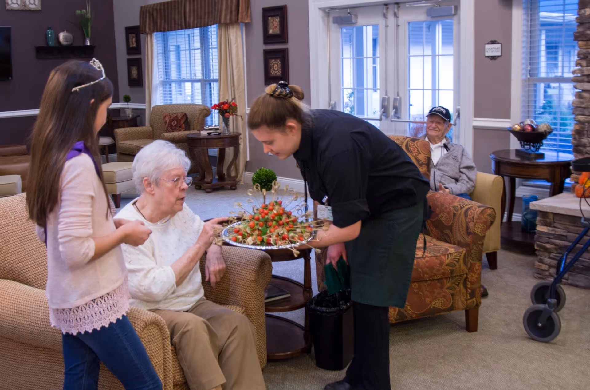 An elderly woman sitting on a couch in a senior living facility common area is being offered a tray of appetizers by a staff member wearing a black shirt and green apron. A young girl stands nearby watching. In the background, an elderly man sits in an armchair near a window and a stone fireplace.