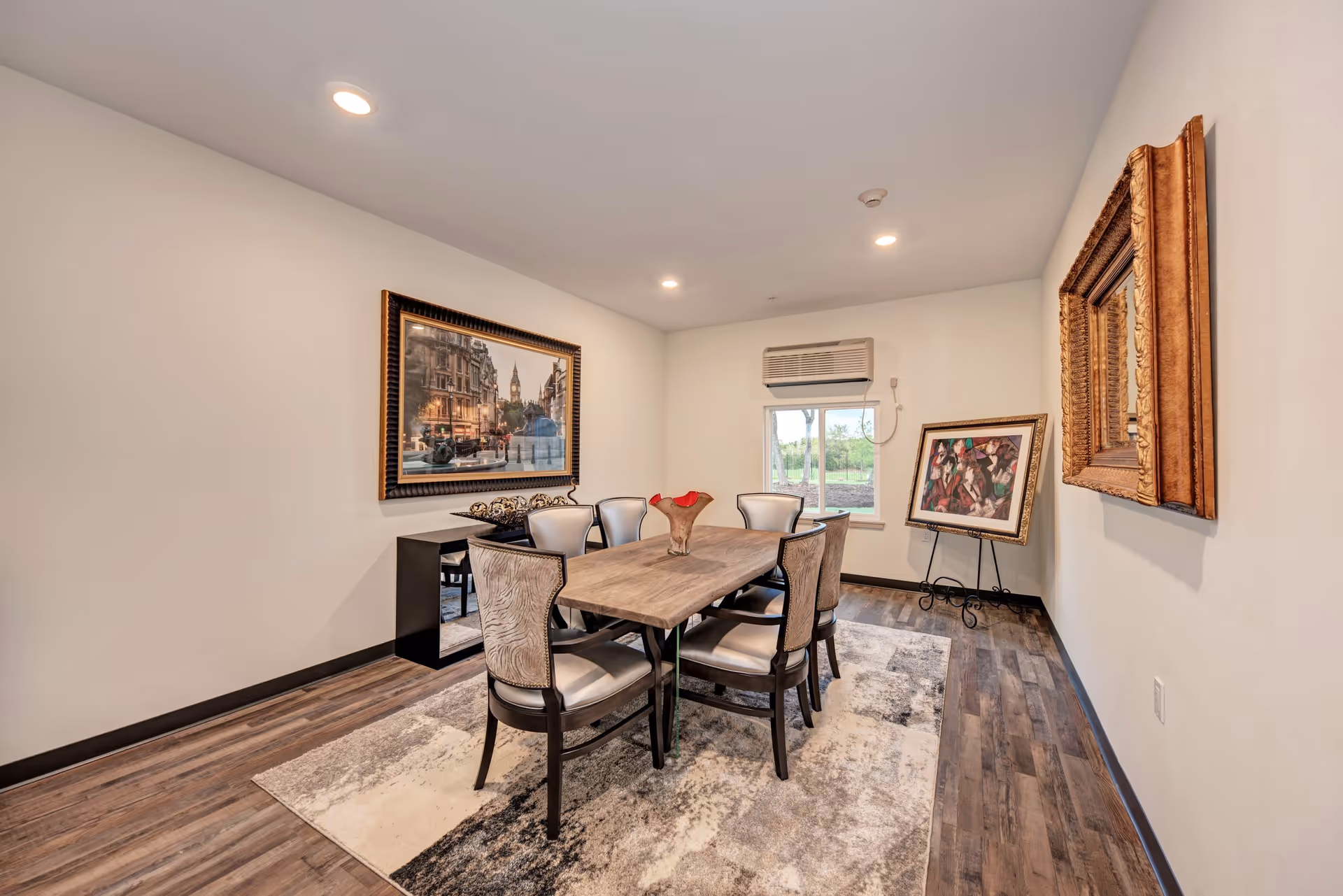 Small dining room with a wooden table and six chairs, framed artwork on the walls, a rug, and a window with a wall-mounted air conditioner.