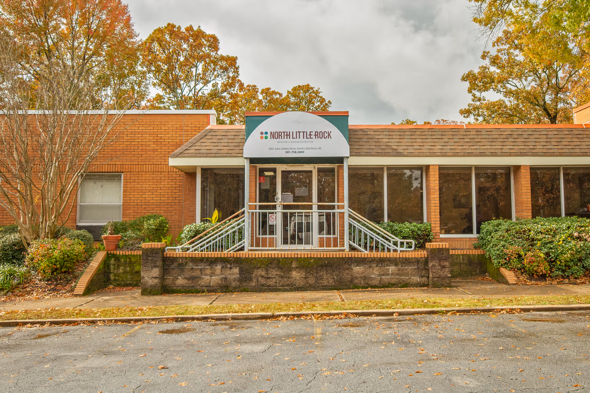 Exterior front view of a single-story brick building with a small entrance porch featuring a ramp and stairs. The building has large windows and is surrounded by bushes and trees with autumn foliage. A sign above the entrance reads 'North Little Rock Health & Rehabilitation'.