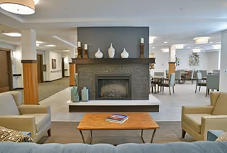 A spacious senior living common area featuring a central fireplace with decorative vases on the mantel. The room includes comfortable armchairs and a wooden coffee table with books, with dining tables and chairs visible in the background under soft ceiling lights.
