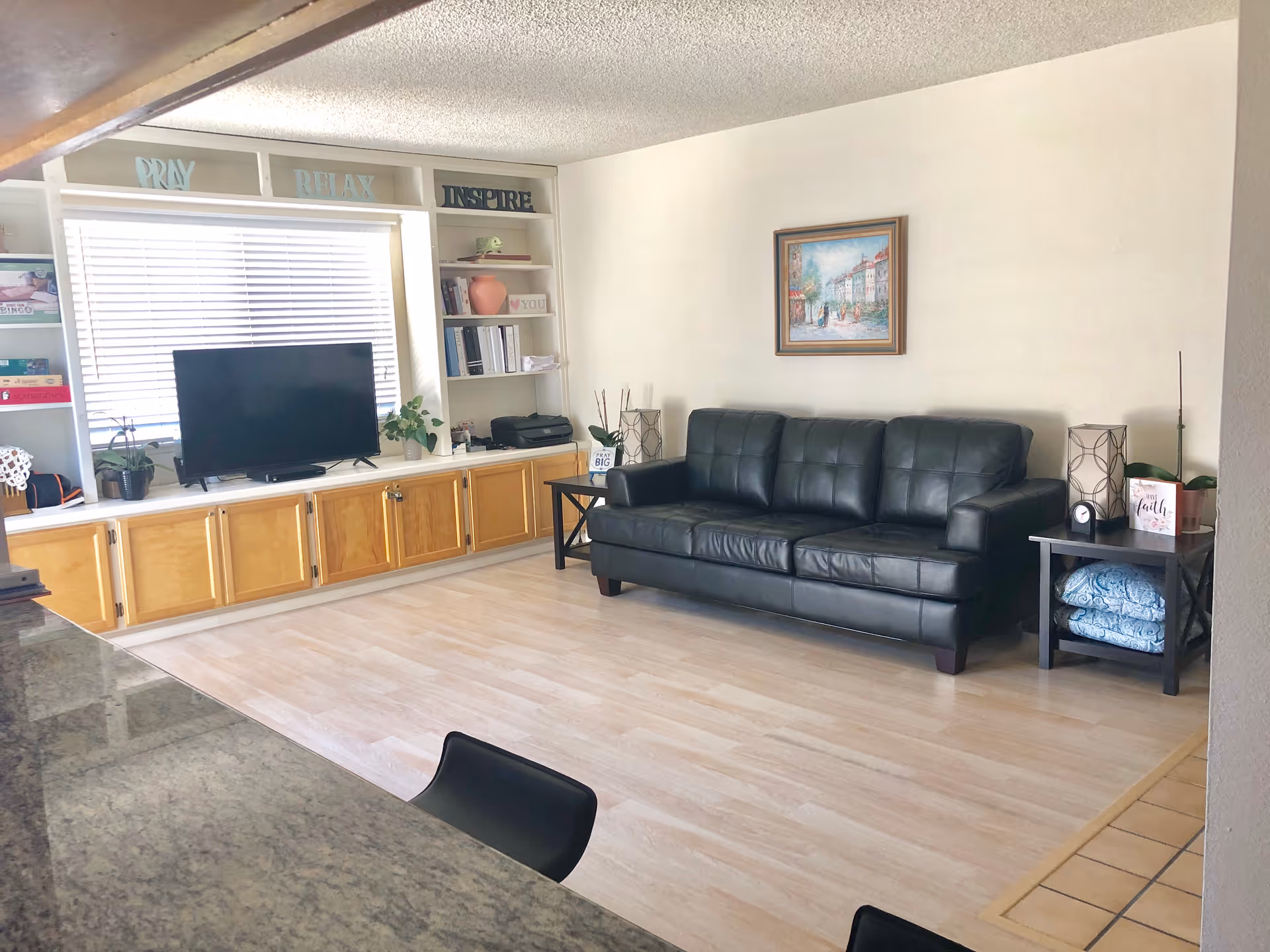 A living room area with a black leather sofa against a white wall, a framed painting above it, and two side tables with lamps and decorative items. Across from the sofa is a built-in wooden cabinet with shelves holding books, plants, and decorative signs that say 'PRAY', 'RELAX', and 'INSPIRE'. A flat-screen TV is placed on the cabinet in front of a window with blinds. The floor is light-colored wood, and part of a granite countertop with black chairs is visible in the foreground.