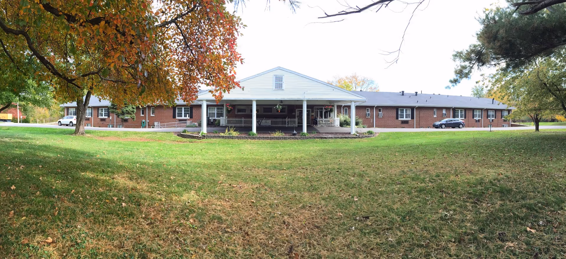 Front view of a one-story brick senior living building with a white columned entrance, surrounded by a grassy lawn and trees with autumn foliage.