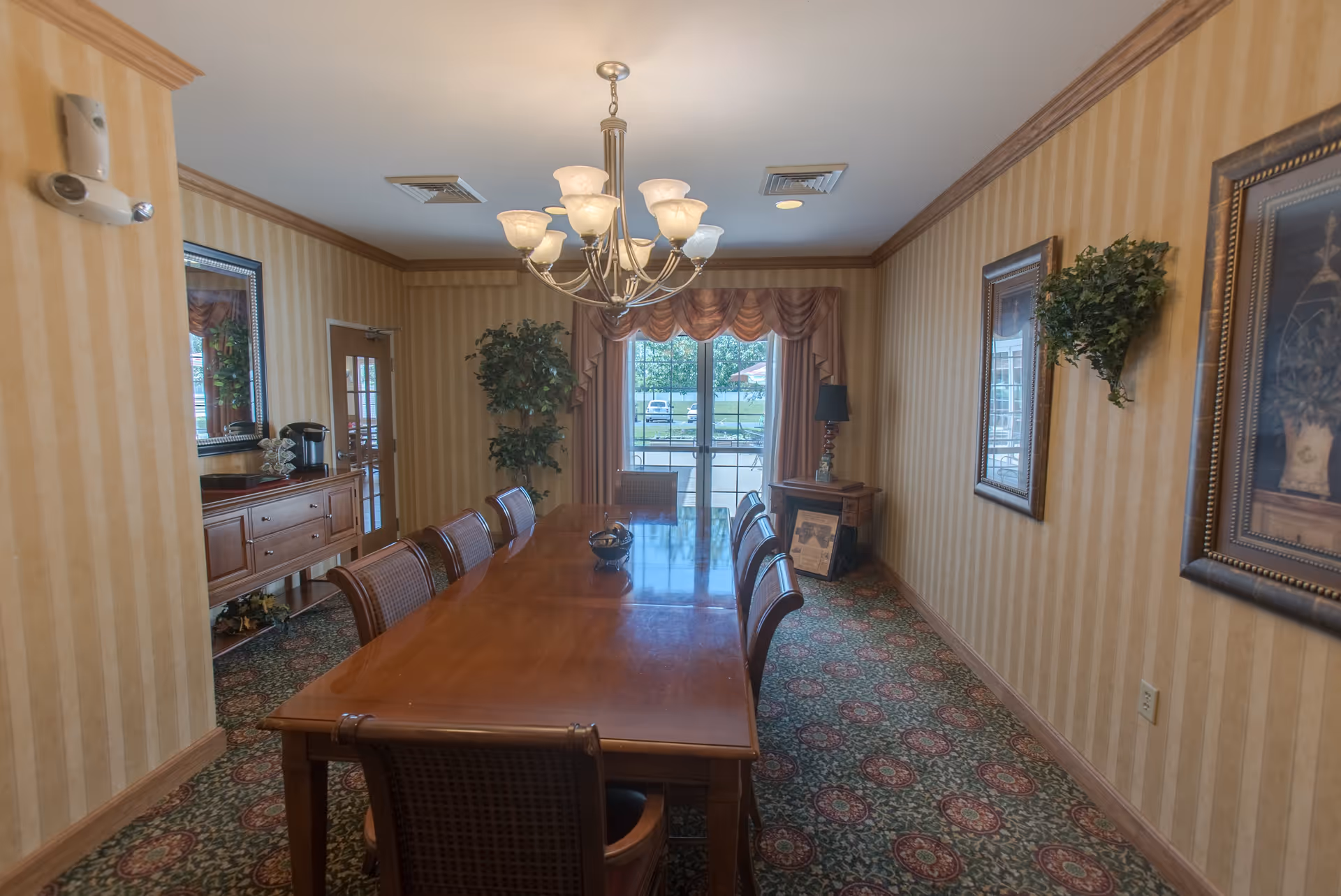 Long formal dining room with a polished wooden table and chairs, chandelier, patterned carpet, framed artwork and a window with drapes.
