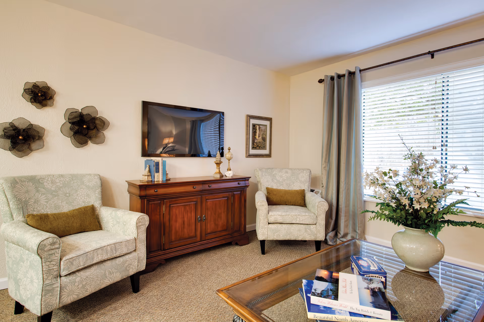 A cozy living room with two light green patterned armchairs each with a brown cushion, a wooden cabinet with decorative items and books, a wall-mounted flat-screen TV, three black decorative wall flowers, a large window with blinds and gray curtains, and a glass coffee table with books and a large vase of white flowers.