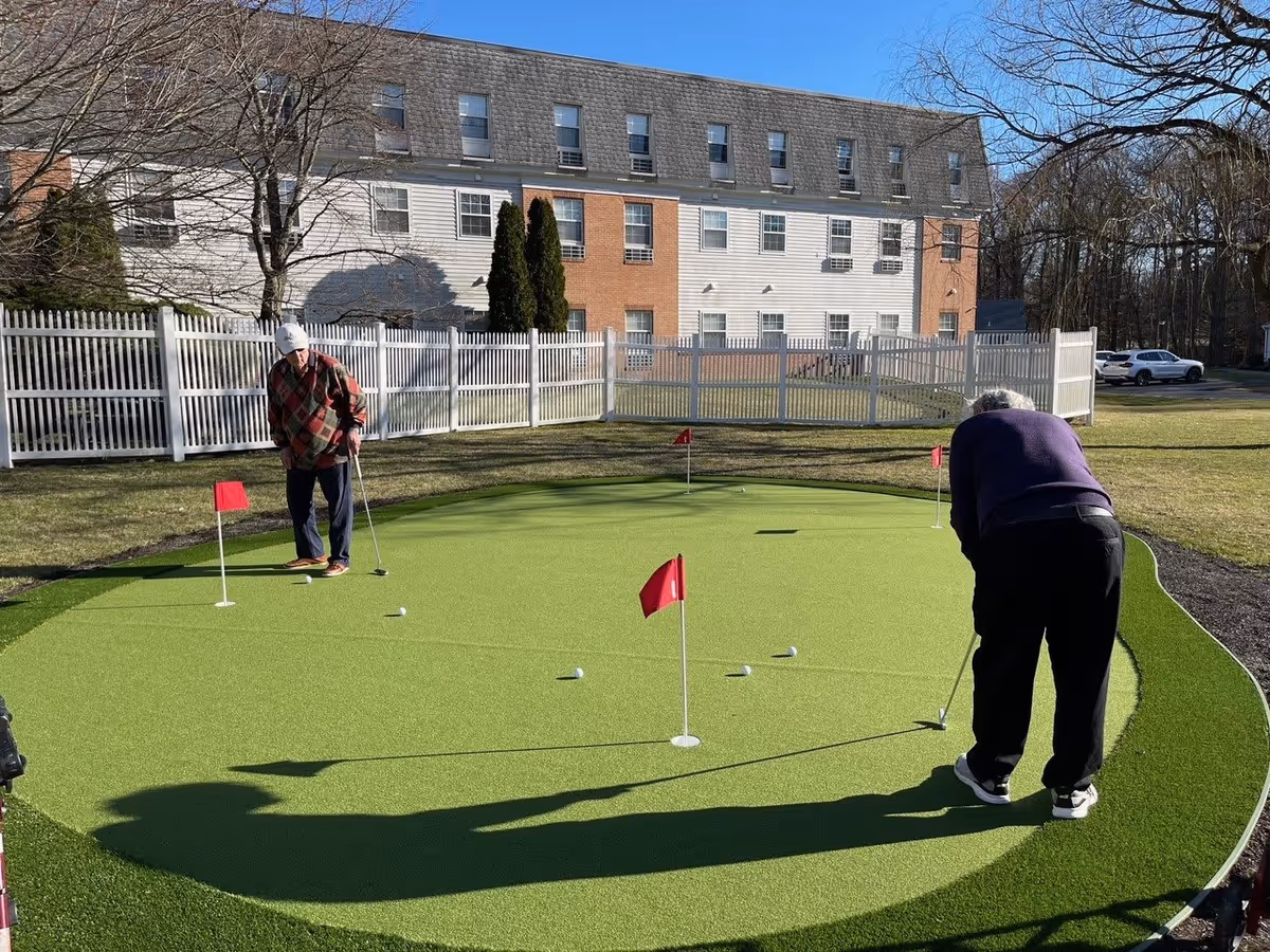 Two elderly men playing golf on a putting green outside, with a white picket fence and a multi-story building in the background under a clear blue sky.