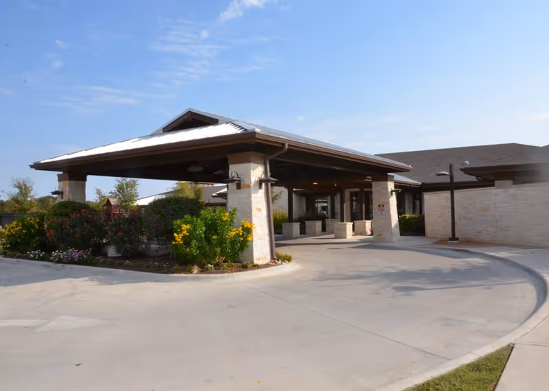 Entrance of a senior living facility with a covered drop-off area supported by stone pillars, surrounded by landscaped bushes and flowers under a clear blue sky.