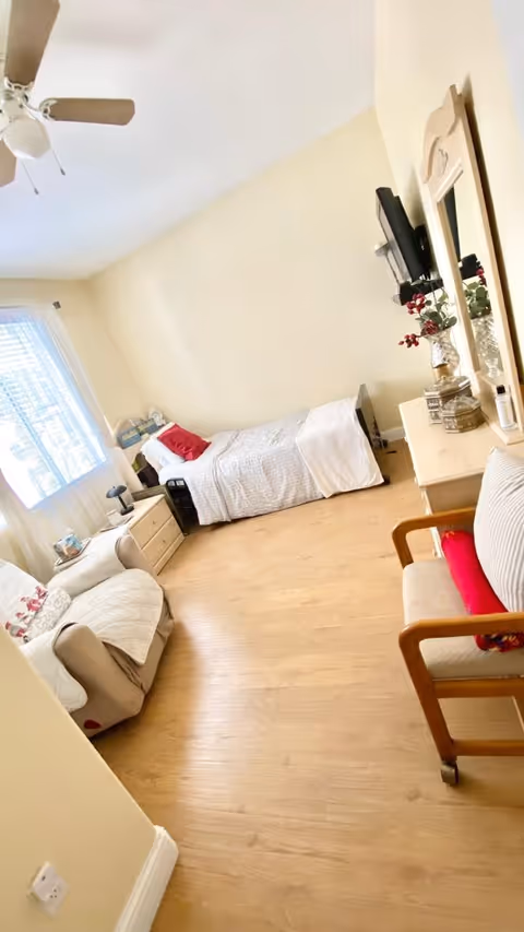 A cozy bedroom with a single bed against the wall, a bedside table with a lamp, a comfortable armchair with a pillow, a wooden chair with a cushion, a dresser with a mirror and decorative items, and a ceiling fan. The room has light-colored walls and wooden flooring, with natural light coming through a window with sheer curtains.