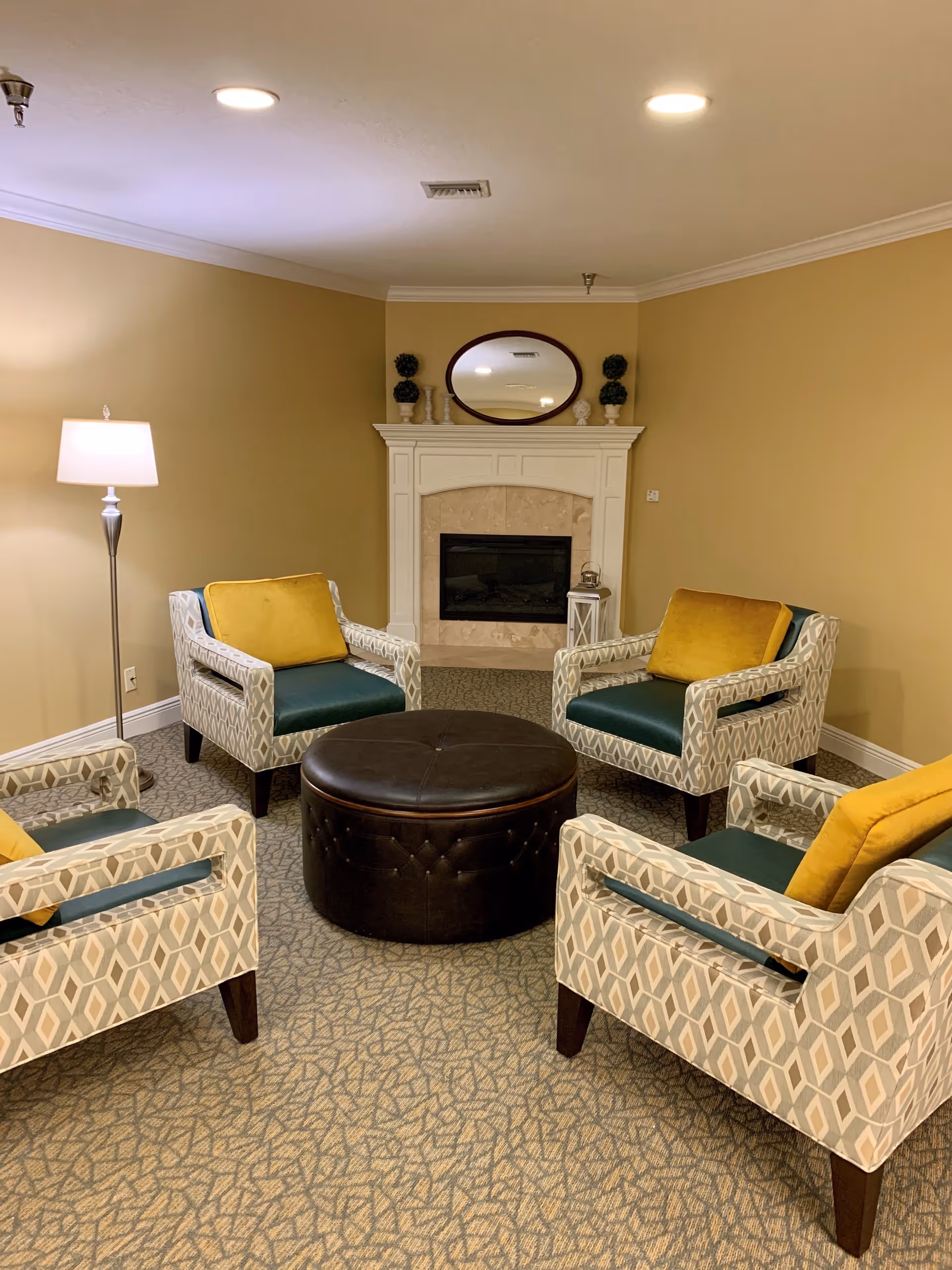A cozy sitting area with four patterned armchairs featuring green seats and yellow cushions arranged around a round, dark brown ottoman. The room has beige walls, a patterned carpet, a white fireplace with a beige tile surround, and a round mirror above the mantel. A floor lamp is positioned in the corner providing warm lighting.