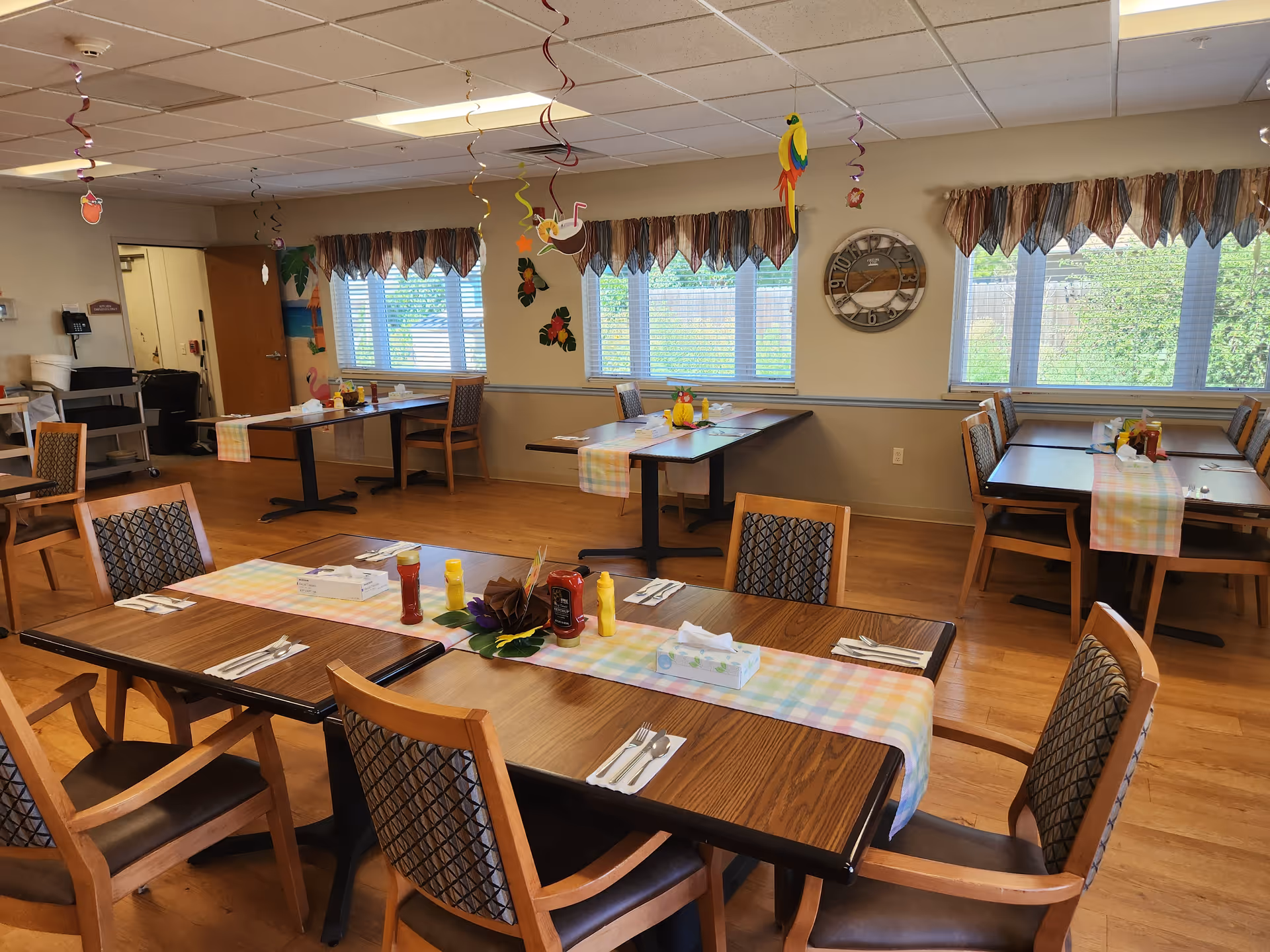 A dining room in a senior living facility with multiple wooden tables and chairs arranged neatly. Each table has a pastel-colored table runner, napkins, utensils, and condiments like ketchup and mustard. The room has large windows with valance curtains, letting in natural light. Colorful hanging decorations, including birds and flowers, are suspended from the ceiling. A large decorative clock is mounted on the wall.