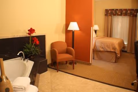 A cozy room in a senior living facility featuring a corner bathtub with towels and a vase, a comfortable armchair, a floor lamp, and a bed with a bedside lamp in the background near a window with curtains.