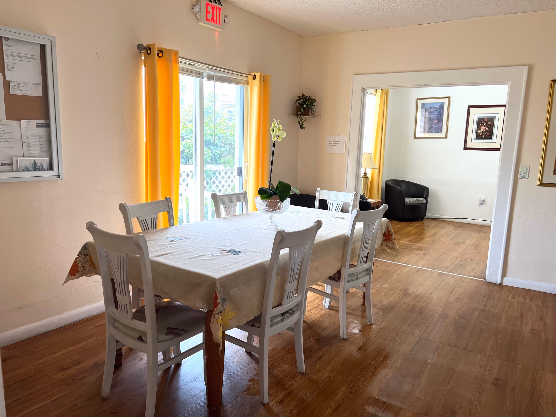 A dining room with a rectangular table covered with a beige tablecloth featuring leaf patterns. Six white wooden chairs surround the table. A potted orchid plant is placed in the center of the table. The room has wooden flooring and cream-colored walls. Bright yellow curtains frame a sliding glass door leading outside. An exit sign is visible above the door. In the adjacent room, a black armchair, a lamp, and framed artwork are visible on the walls.