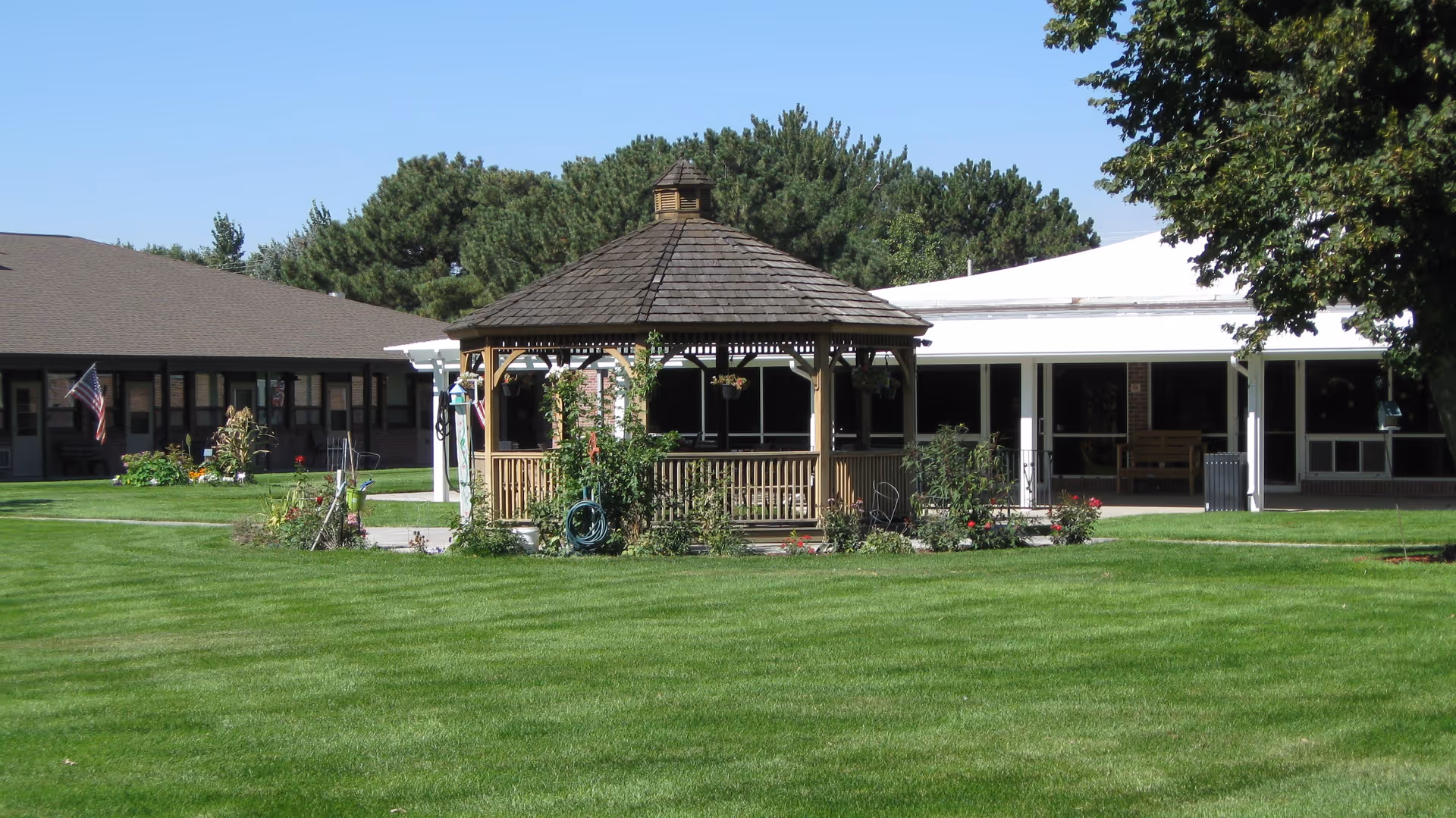 A wooden gazebo with a shingled roof sits in the middle of a well-maintained green lawn with flower beds around it. Behind the gazebo, there are single-story buildings with large windows and a covered porch area. Trees and shrubs surround the area under a clear blue sky.