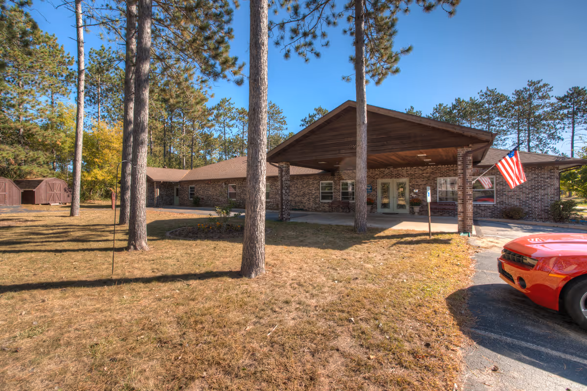Exterior view of a single-story brick building with a covered entrance supported by brick columns. There are tall pine trees in the front yard, an American flag near the entrance, and a red car parked on the driveway. The sky is clear and blue.