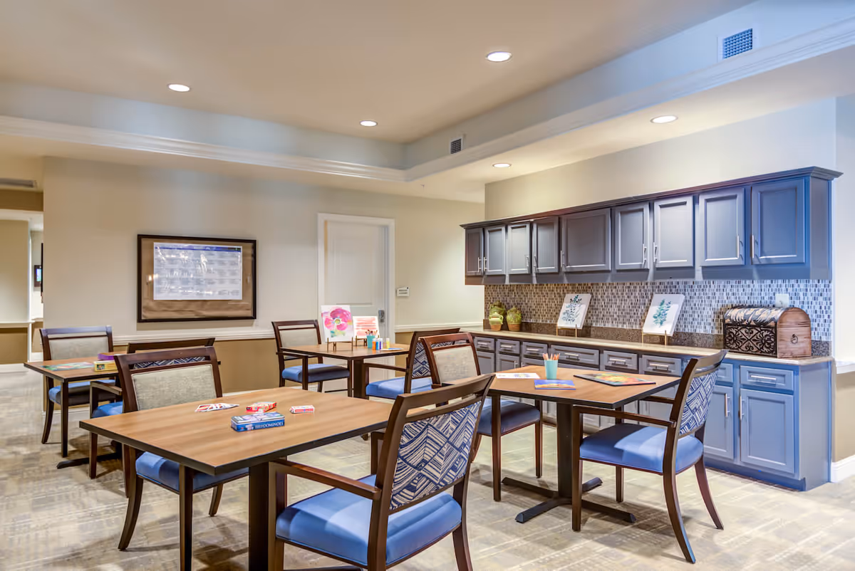 A bright and clean activity room with several wooden tables and chairs arranged for group activities. The tables have art supplies, coloring books, and card games on them. The room features blue cabinets with a mosaic tile backsplash along one wall, and framed artwork displayed on the countertop. The walls are painted a light beige color, and the ceiling has recessed lighting.