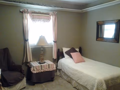 A small bedroom with a single bed covered in a white quilt and two pillows, one brown and one pink. Next to the bed is a nightstand with a striped tablecloth and a lamp. There is a window with pink curtains behind the nightstand. A brown armchair is partially visible on the left side of the image. A rectangular mirror with a wooden frame hangs on the wall above the bed.
