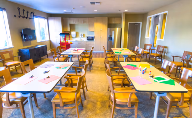 A bright room with several tables and wooden chairs arranged for an activity or craft session. The tables have colorful paper, glue sticks, scissors, and other craft supplies. The room has large windows letting in natural light, a popcorn machine in the back, and a kitchenette area with cabinets and a refrigerator.