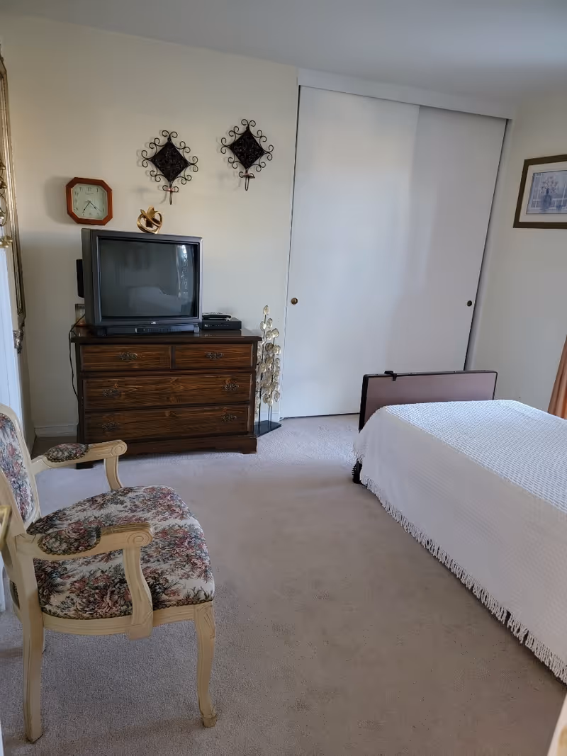 Bedroom with a bed covered by a white bedspread, a floral upholstered armchair, and a wooden dresser topped with a TV in front of sliding closet doors.