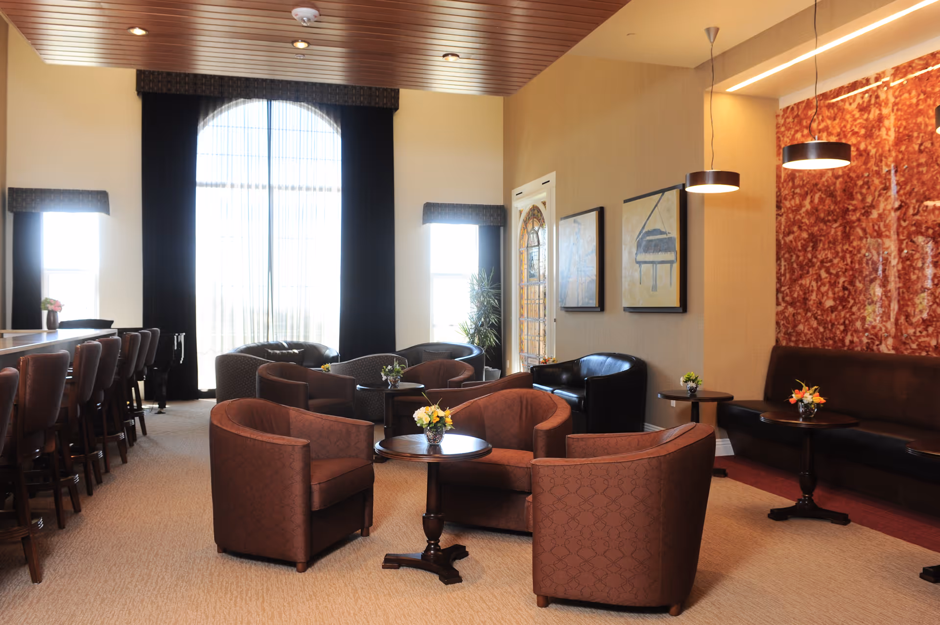 A spacious and well-lit lounge area with multiple brown upholstered armchairs arranged around small round tables with flower centerpieces. There is a long bar with high chairs on the left side, large windows with sheer curtains letting in natural light, and modern pendant lights hanging from the ceiling. The walls feature framed artwork and a decorative red panel.