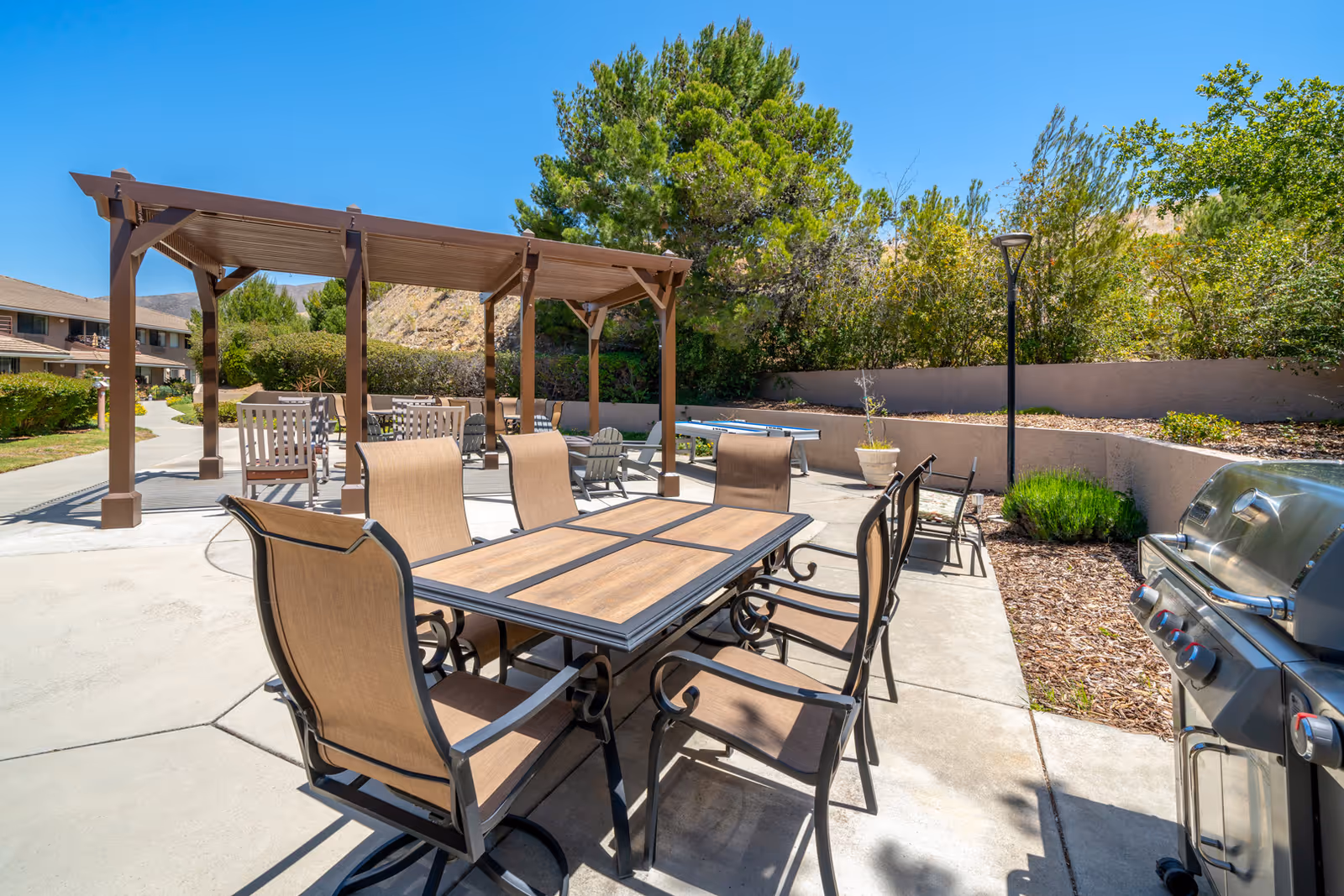 Outdoor patio area at Las Brisas Retirement Community featuring a large dining table with six chairs, a barbecue grill, a shaded pergola with additional seating, and surrounding greenery under a clear blue sky.