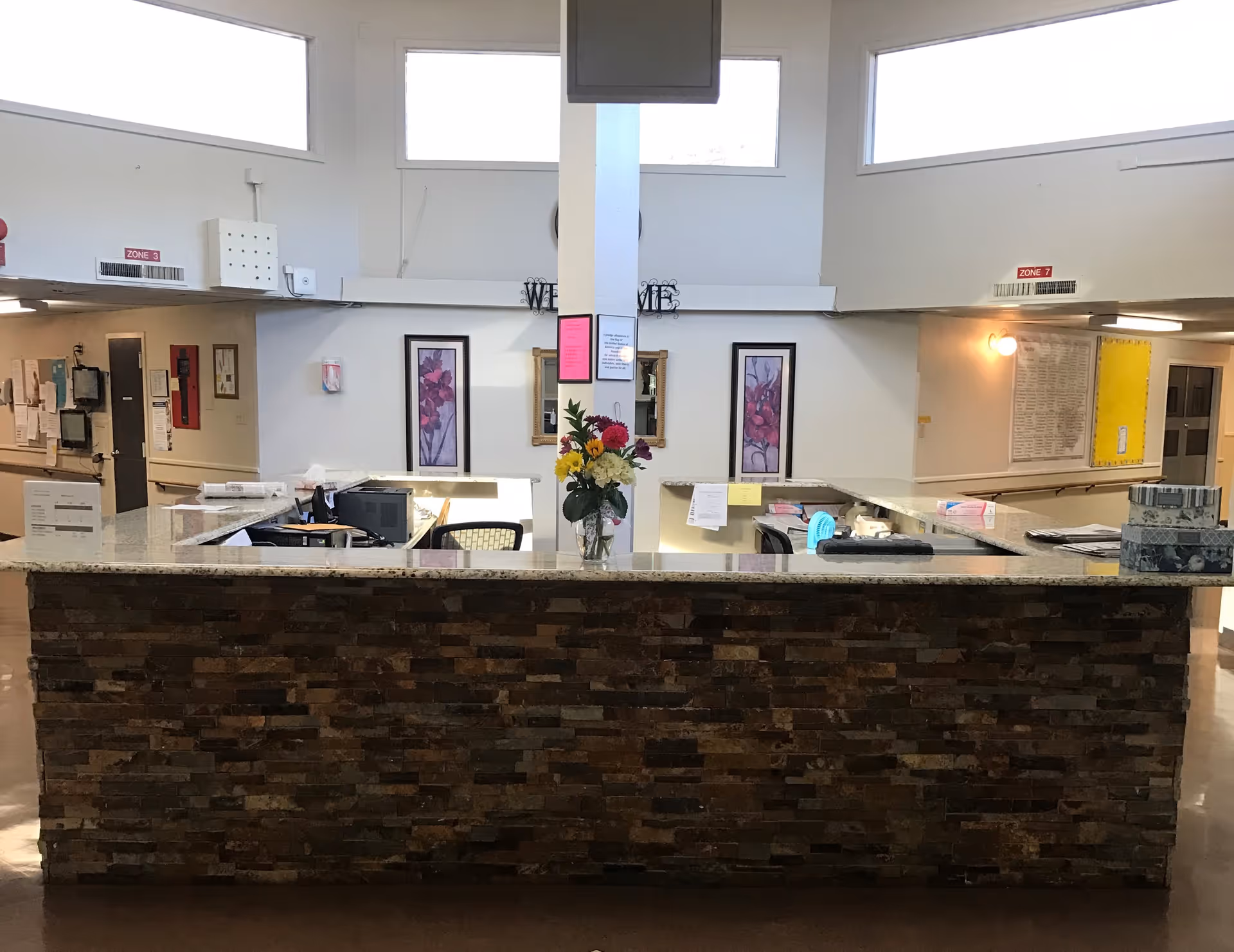 Reception desk area inside a nursing and rehabilitation center with a stone front counter, a vase with colorful flowers on the counter, office chairs, computer monitors, and framed floral artwork on the wall behind. The area is well-lit with high windows and has signs indicating zones on the walls.