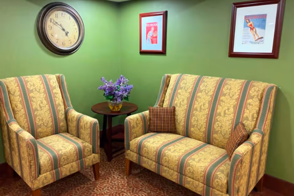 A cozy sitting area with a patterned armchair and matching loveseat featuring yellow and green stripes with floral designs. Between them is a small round wooden table holding a vase of purple flowers. The walls are painted green and decorated with a round clock and two framed vintage-style posters.