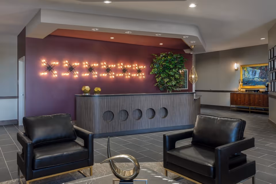 Modern reception area with a wooden front desk featuring circular cutouts, a purple accent wall with decorative lighting, two black leather armchairs, a round marble table with a metallic sculpture, a large green plant, and a sideboard with a framed painting in the background.
