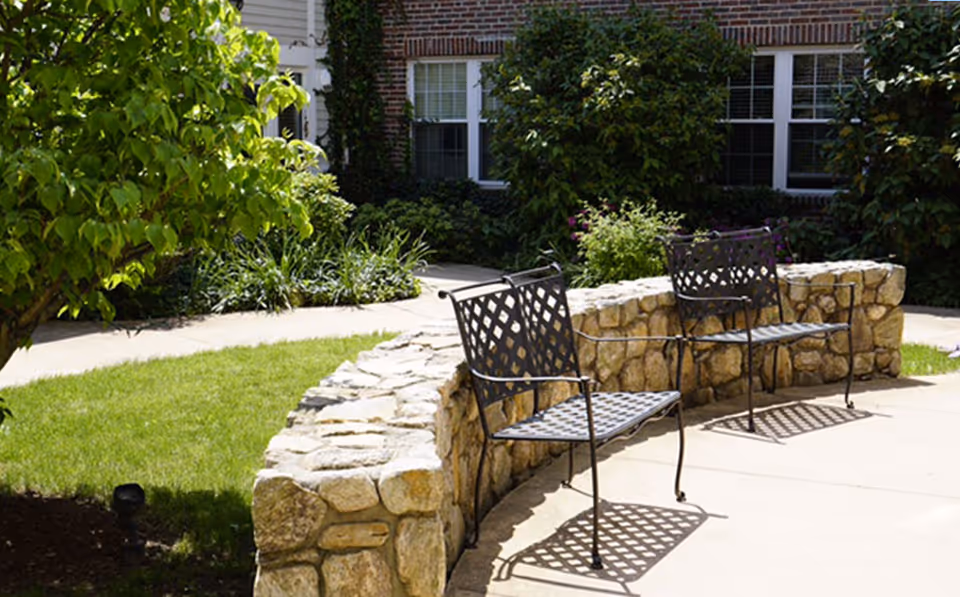 Outdoor seating area with two black metal benches placed on a curved stone wall in a garden with green grass, shrubs, and a tree near a brick building with windows.