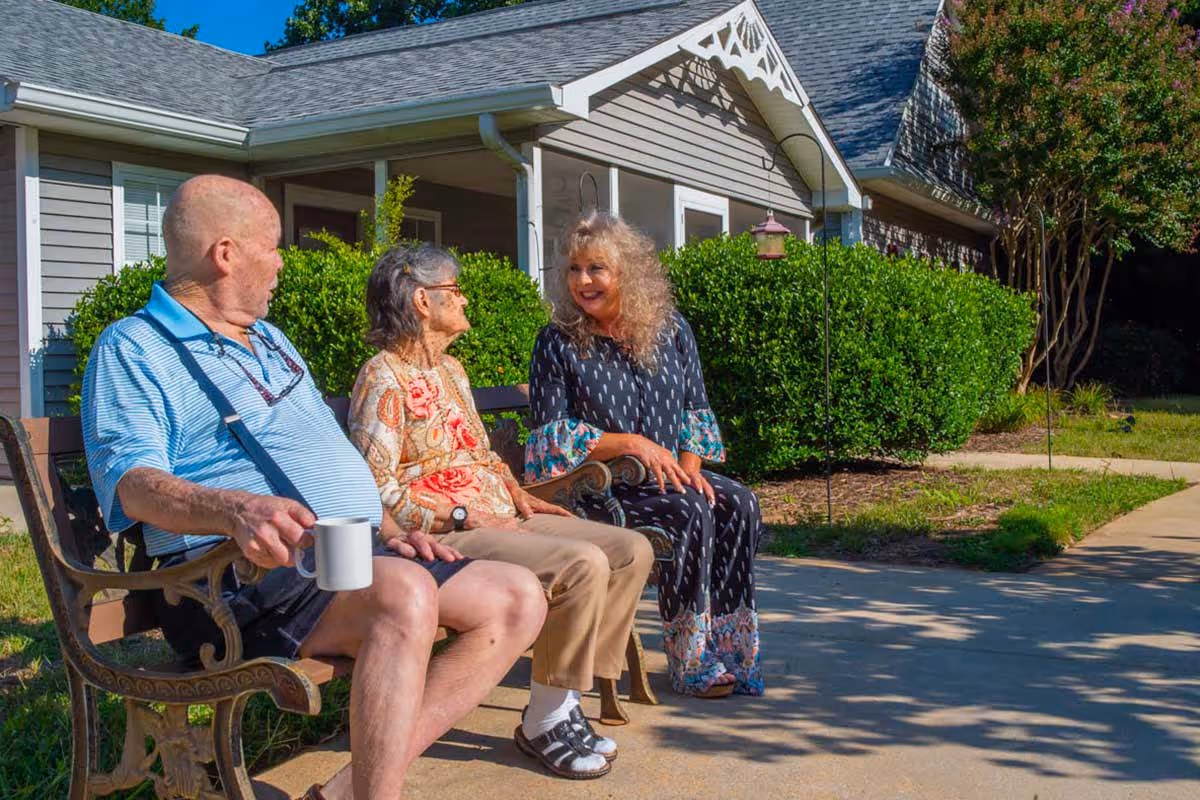 Three elderly people sitting on a bench outside a residential building, enjoying a sunny day. One man is holding a white mug, and two women are engaged in conversation. The background shows green bushes and part of the building with a gray roof and siding.