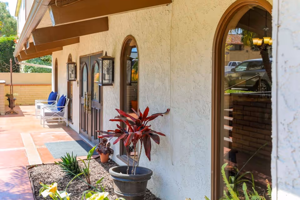 Exterior view of Sea Cliff Assisted Living showing a walkway with potted plants and white chairs with blue cushions along a stucco wall with arched windows and a double wooden door under a brown awning.