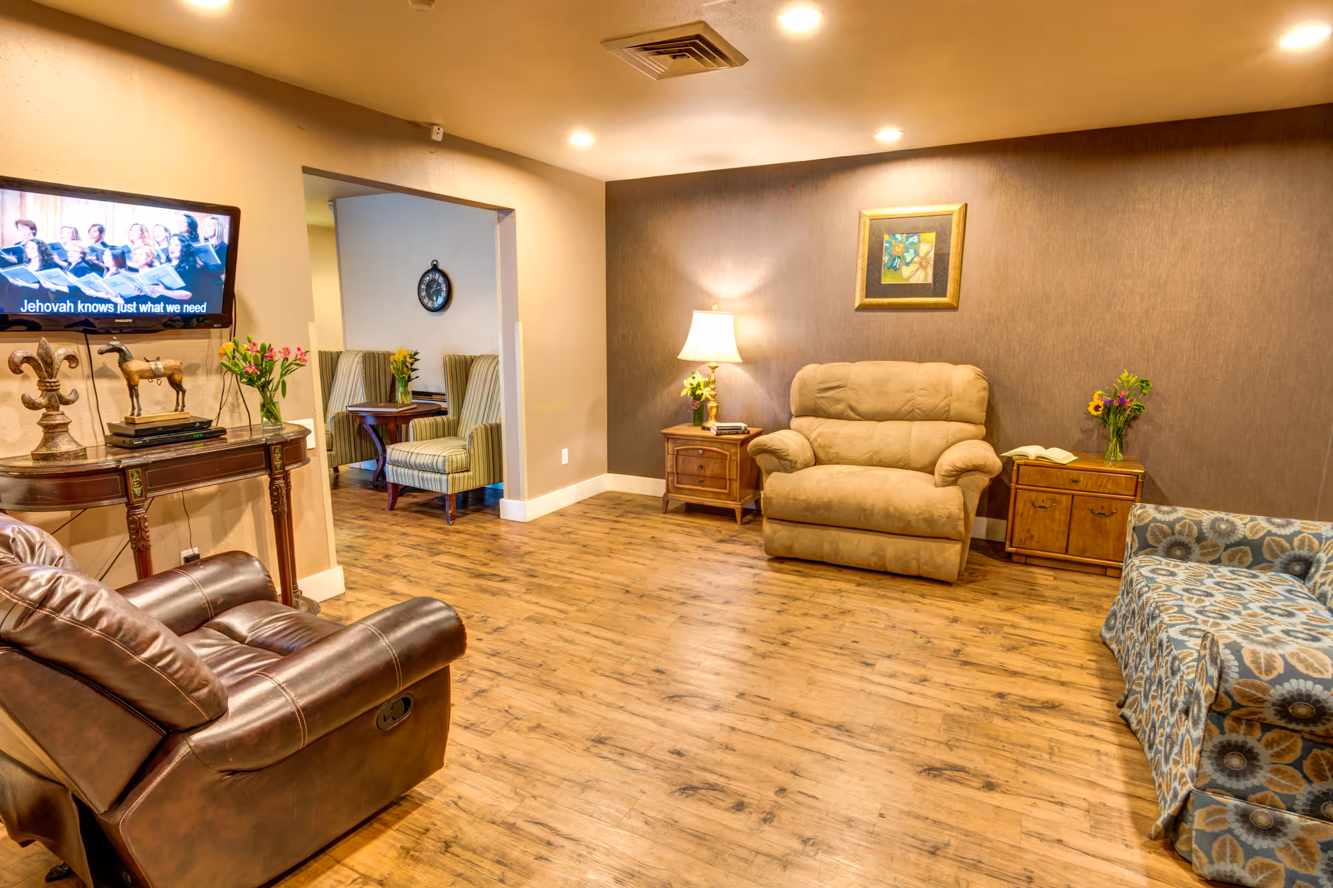 A warm assisted-living lounge with recliners and sofas arranged on wood floors, a wall-mounted TV, side tables, and lamps.