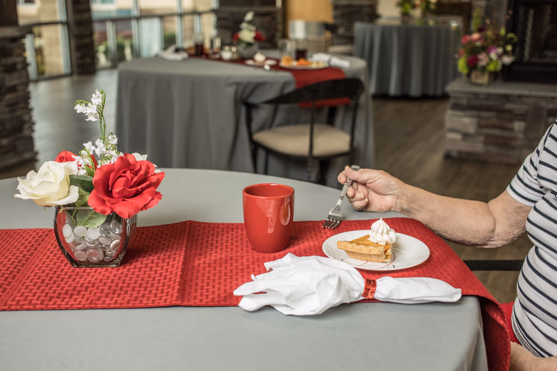 A close-up of a dining table with a red table runner, a glass vase with red and white flowers, a red mug, and a plate with a slice of pie topped with whipped cream. An elderly person wearing a striped shirt is holding a fork poised to eat the pie. In the background, there are more tables with gray tablecloths and chairs in a well-lit dining area.
