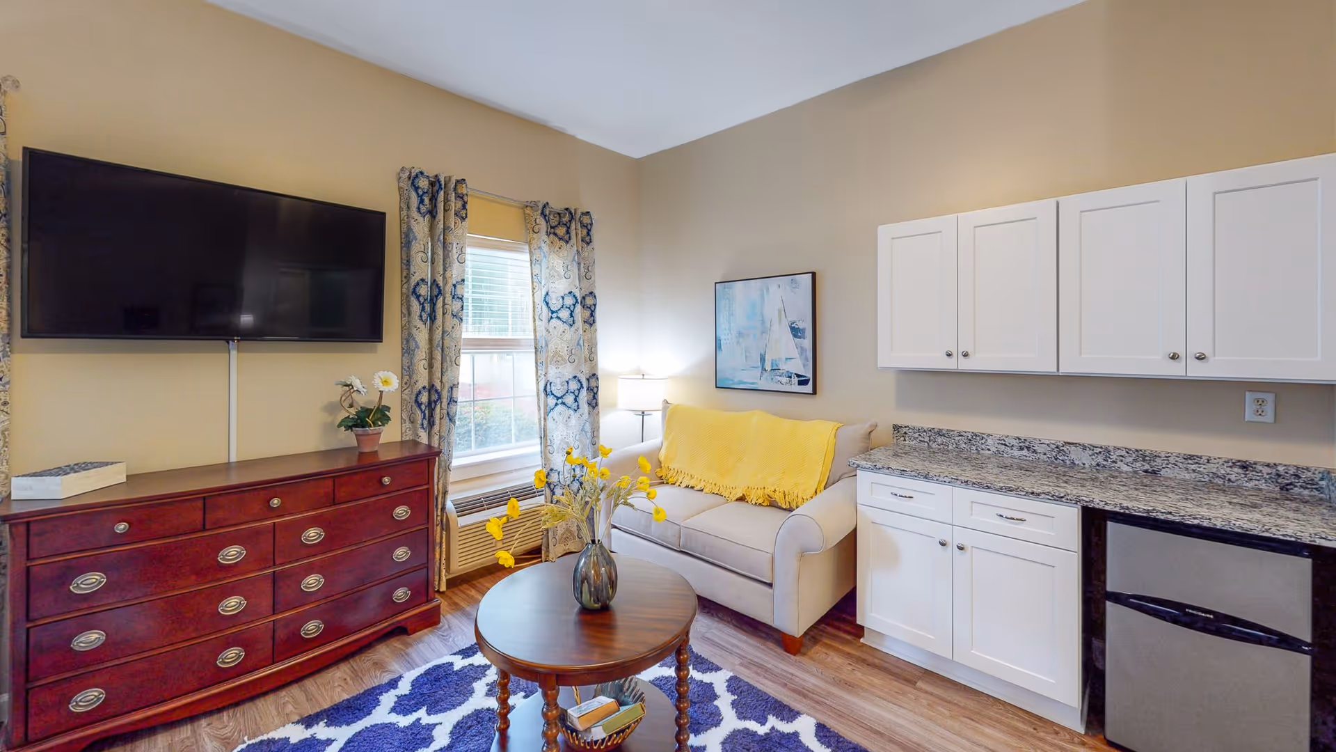 A cozy living room area featuring a beige loveseat with a yellow throw blanket, a round wooden coffee table with a vase of yellow flowers, a large flat-screen TV mounted on the wall above a wooden dresser, patterned curtains on a window, white cabinets with granite countertop, and a small refrigerator underneath the counter.