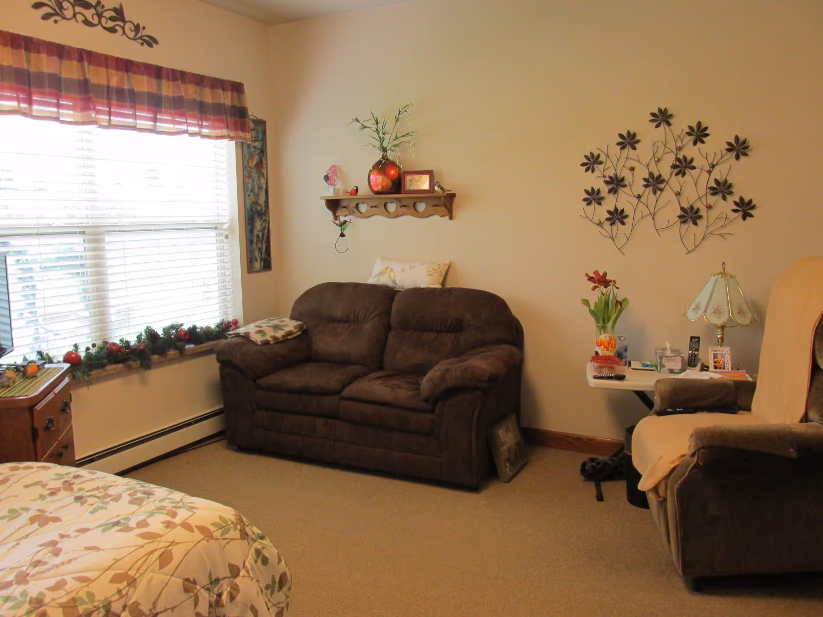 Cozy assisted-living room with a brown loveseat and recliner, floral wall art, a window with blinds and valance, and a small side table with a lamp.