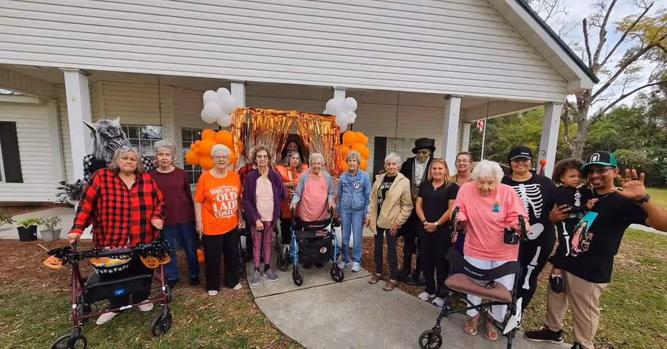 A group of elderly people and caregivers standing outside a building decorated with orange and white balloons and orange streamers. Some individuals are using walkers, and a few are dressed in Halloween-themed costumes, including a werewolf mask and skeleton outfit. The setting appears festive and cheerful.