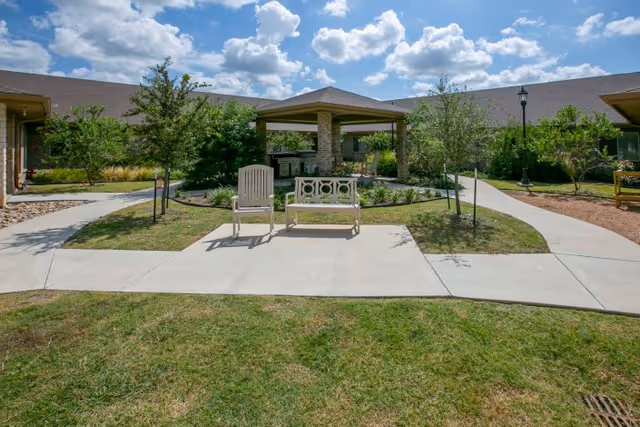 Sunny courtyard with a gazebo, a bench and a chair surrounded by walkways and landscaping in front of a single-story building.
