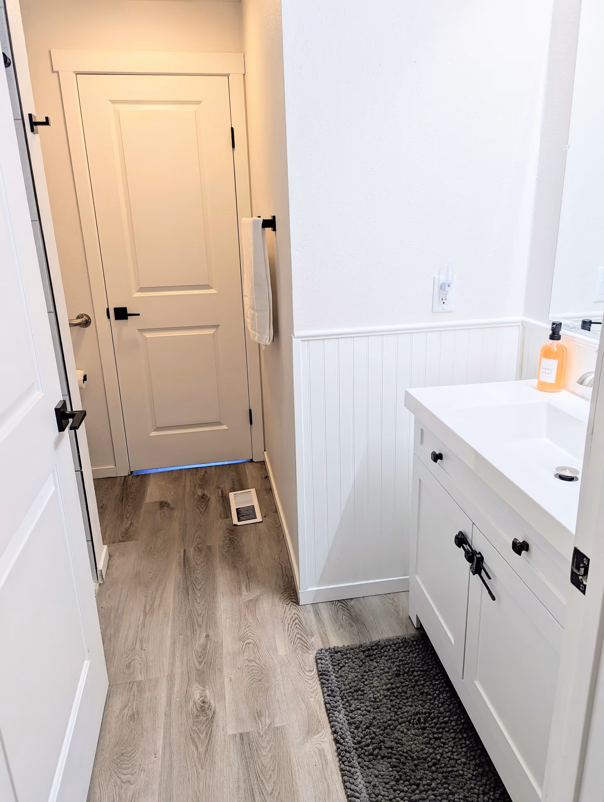 A clean and modern bathroom with light wood flooring, white walls, and a white vanity with a sink. There is a dark gray textured bath mat in front of the vanity. A towel hangs on a black towel rack on the wall near a closed white door. A bottle of hand soap is placed on the vanity next to the sink.