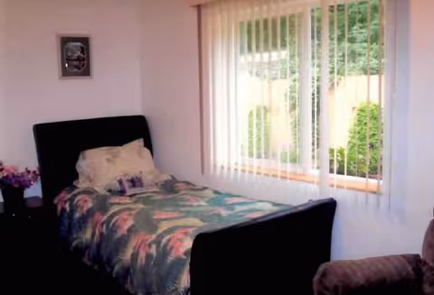 Bedroom with a single bed, floral quilt, pillows, and a window with vertical blinds.